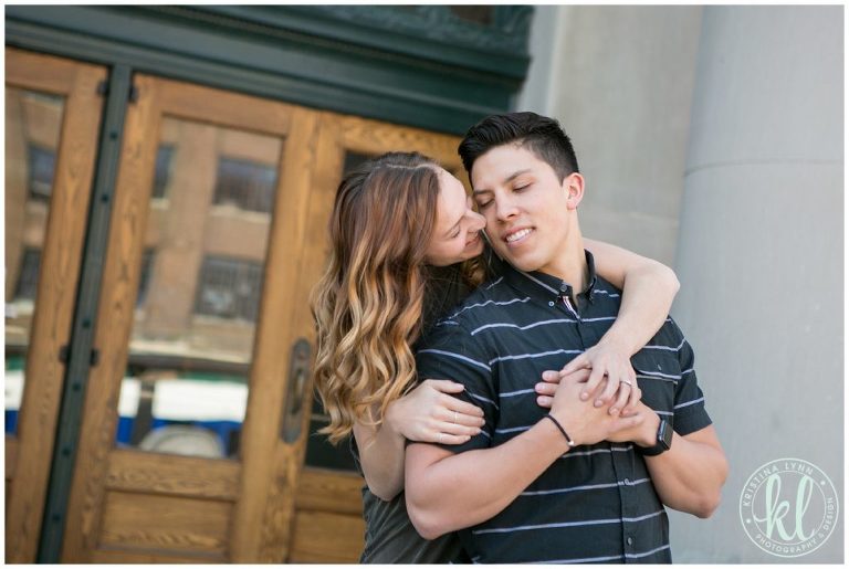 Girl wraps her arms around her guy and kisses him on the cheek.