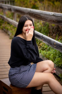 Kristina Lynn Photography & Design Image of high school senior girl dressed in a black top and boots and a checkered skirt, laughs towards the camera while seated outside on a bridge. Senior pictures you'll love in MN, IA, CO, FL.