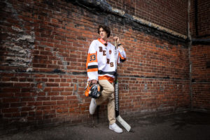 Kristina Lynn Photography & Design Image of high school senior male posing against a brick wall wearing dress pants and his hockey uniform. Holding his other gear. Sporty, outdoor, senior pic ideas.