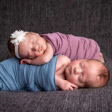 Two newborn babies, one wrapped in blue and the other wrapped in purple wearing a pink hairband and bow snuggle sleepily for an in home photo shoot by Kristina Lynn Photography & Design