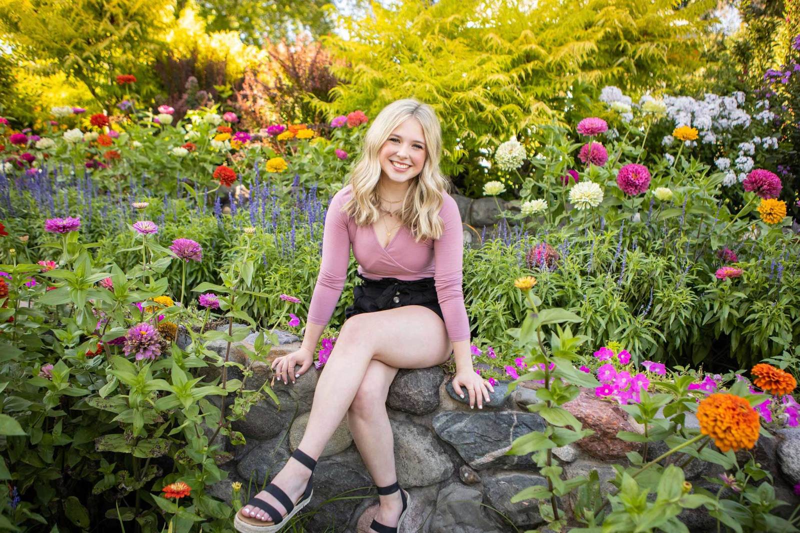 Kristina Lynn Photo of High School Senior girl sitting on a rock wall surrounded by tall colorful flowers