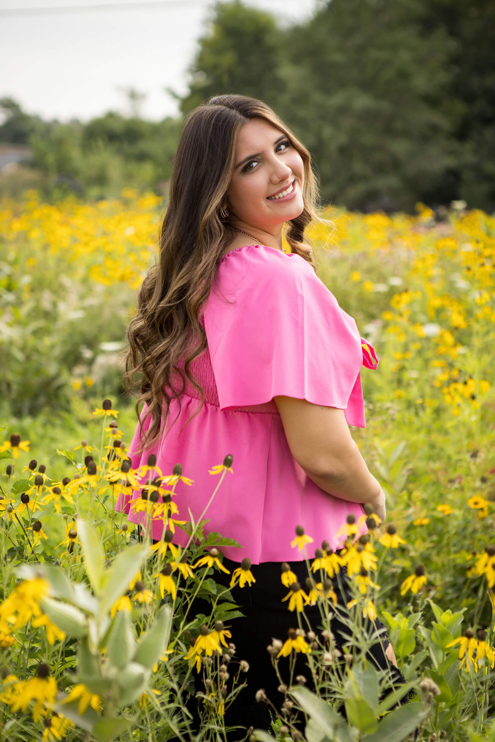 A high school senior portrait of a young woman in a bright pink shirt standing in a field of Black Eyed Susan flowers. The yellow petals set off her outfit nicely to make the image pop.
