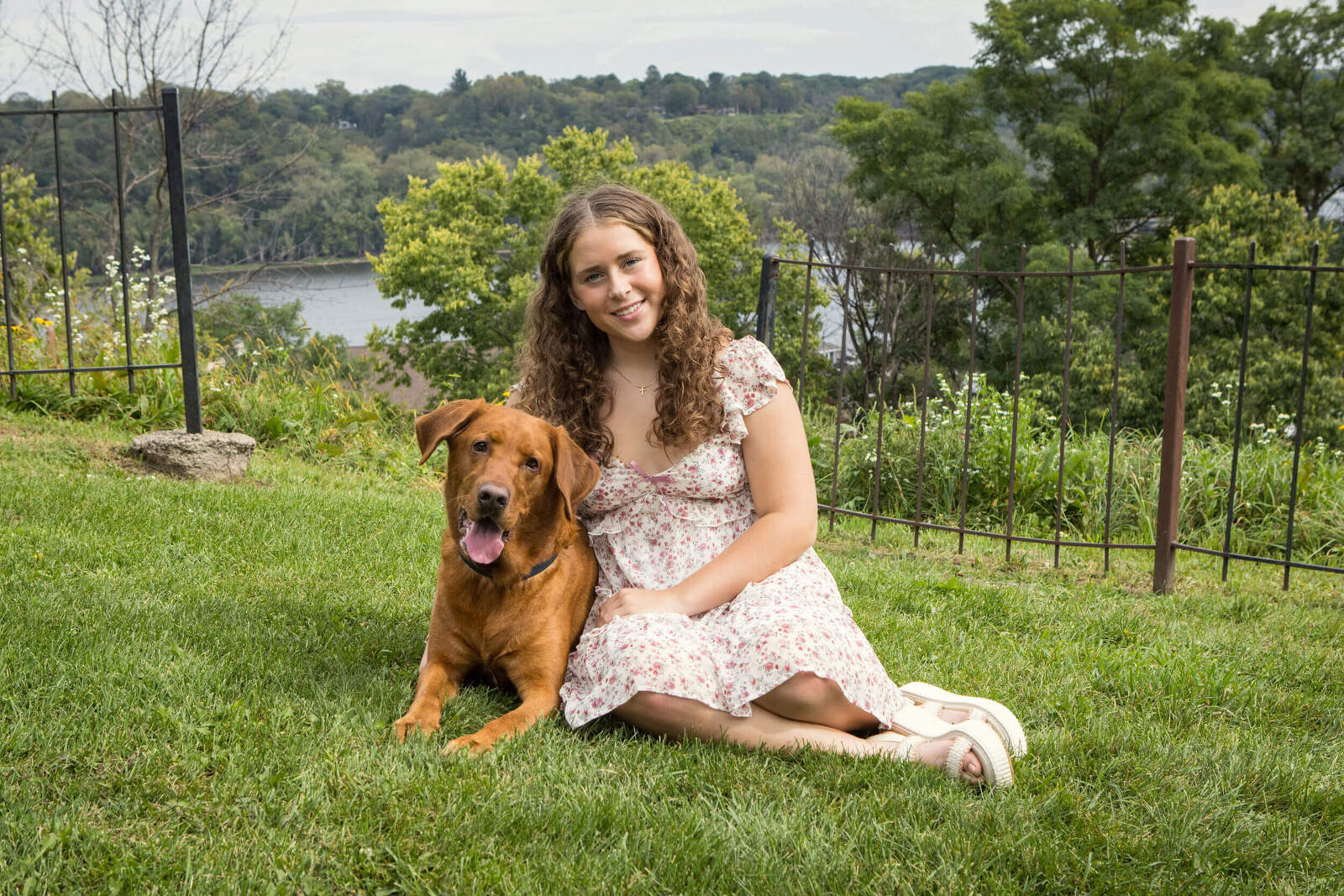 A senior picture taken by professional award winning photographer Kristina Lynn Marshall. The photo features a young woman and her dog, seated in the grass together. The river makes a breathtaking backdrop