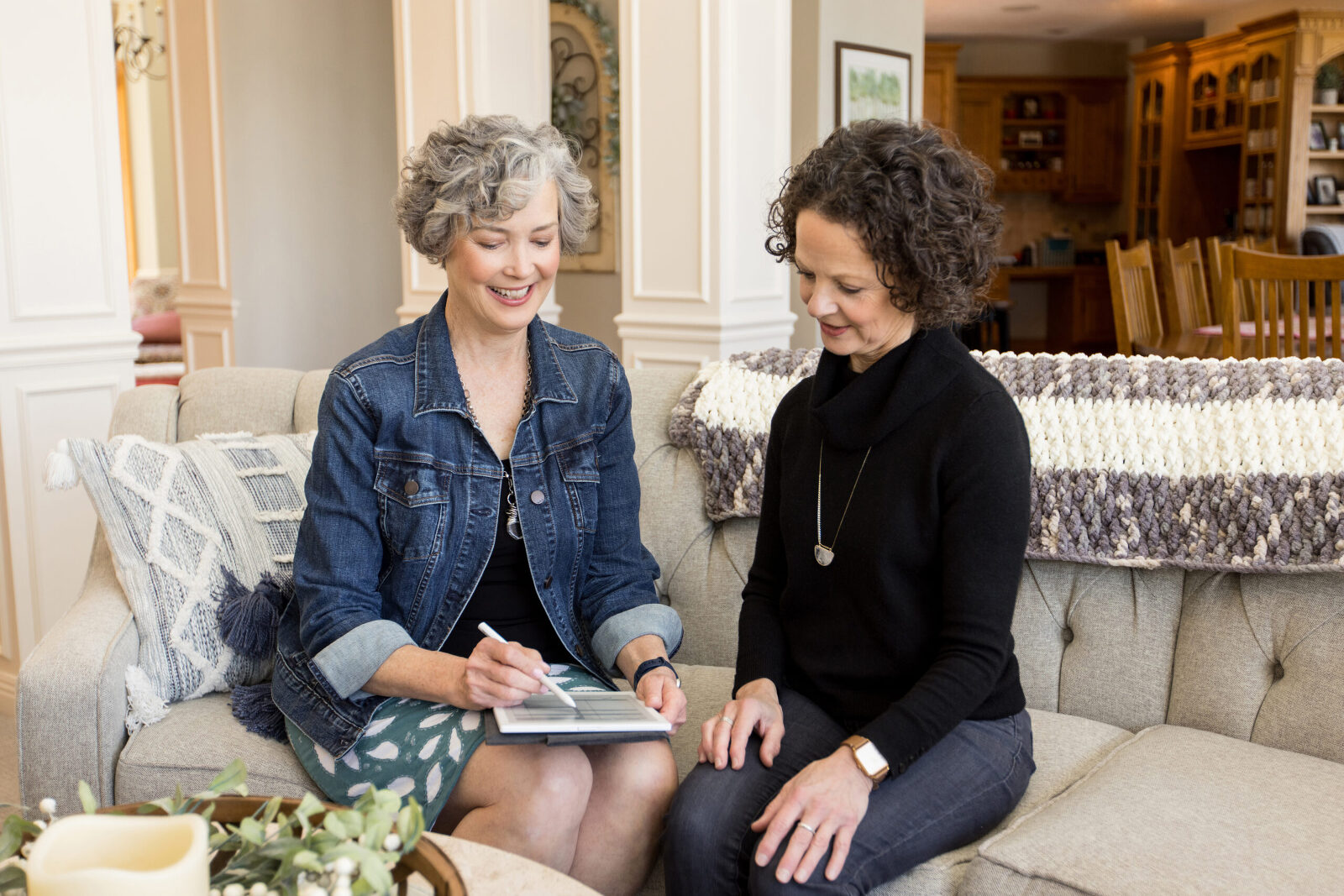 Two women dressed in business casual attire sit on a living room sofa together, looking over a notebook together.