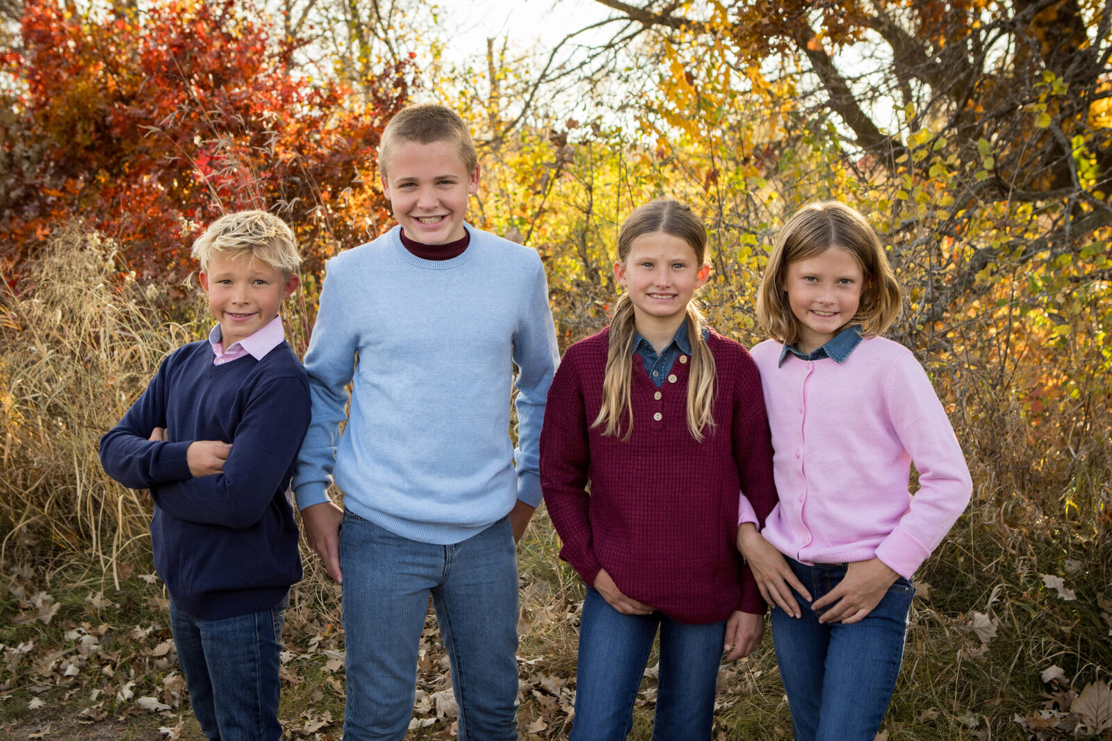 Four siblings (two boys and two girls) in coordinating outfits stand in the fall foliage together, smiling at the camera.