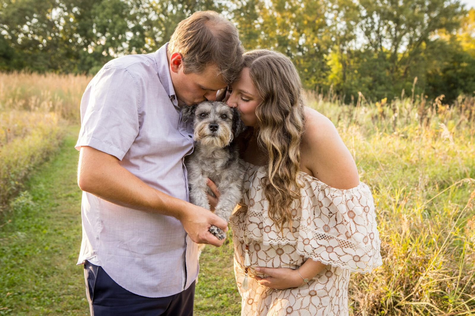 A professional photo of an expecting mother and her male partner snuggling a dog between the two of them. They are both kissing the little grey dogs head while it looks at the camera.