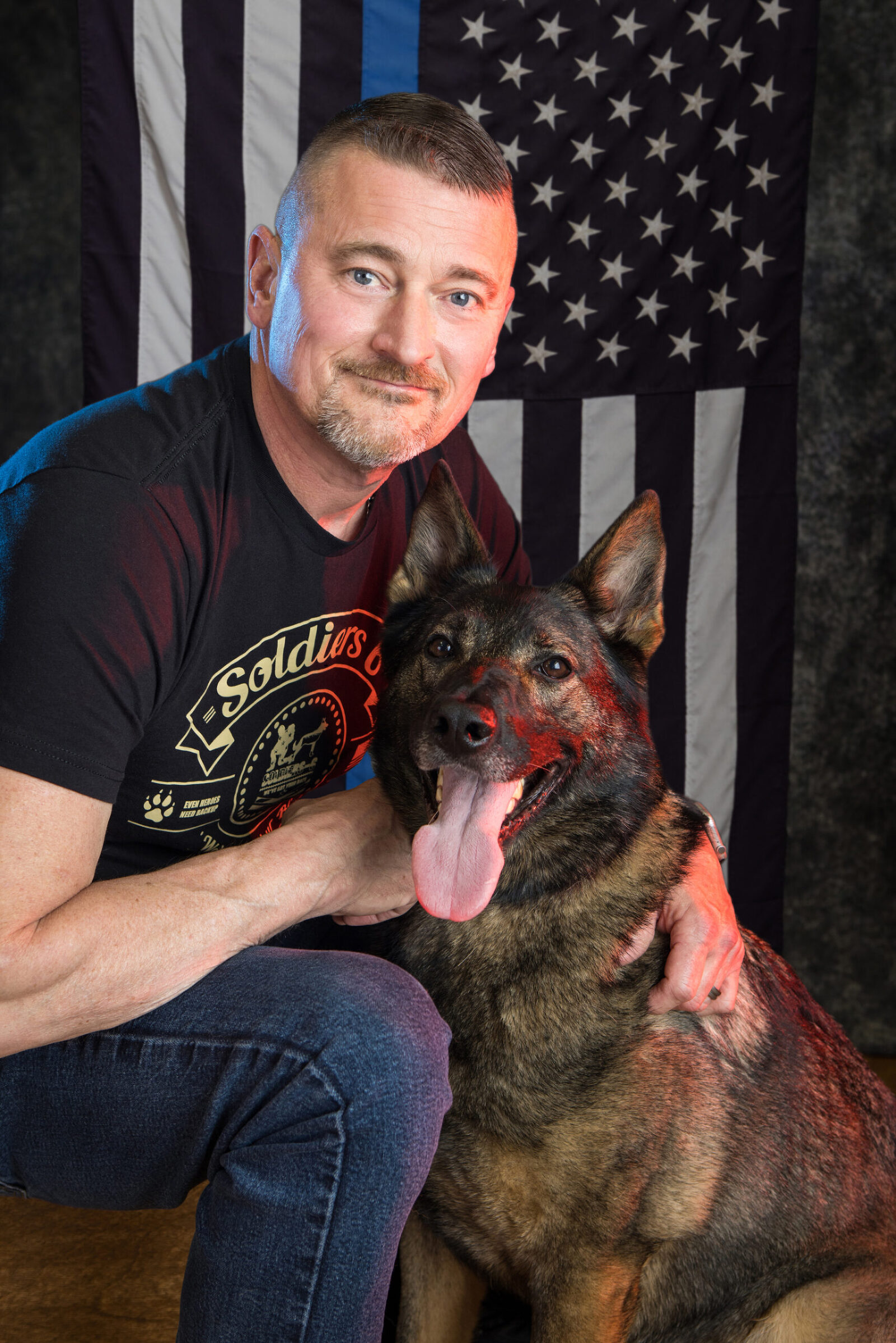 A studio portrait of a man and a German shepherd dog. They are seated in front of a thin blue line American flag.