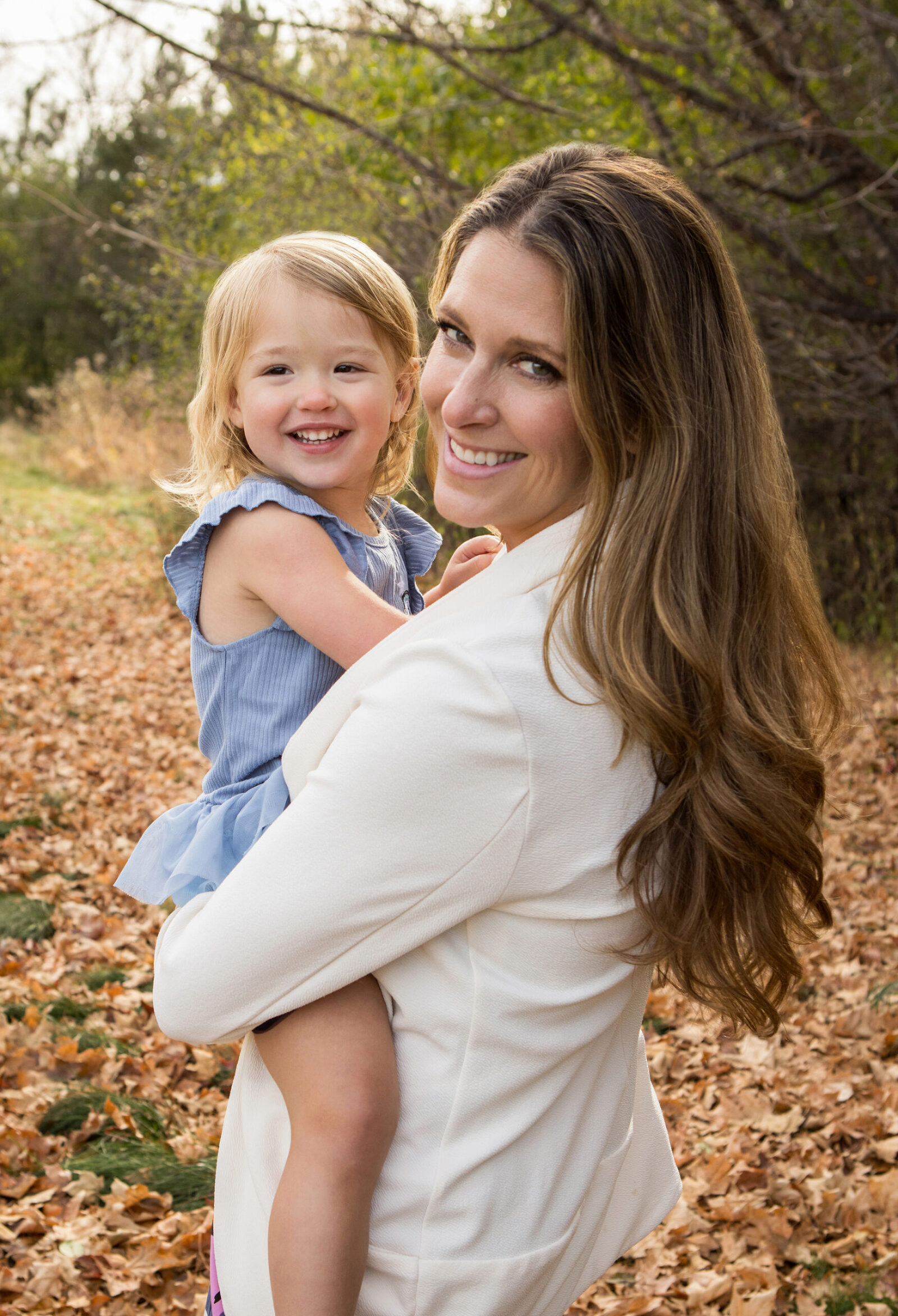 A mother dressed in white looks back over her shoulder smiling at the camera. In her arms is her daughter, dressed in blue smiling while looking off to the side. The setting is in a park, with fall leaves on the ground.