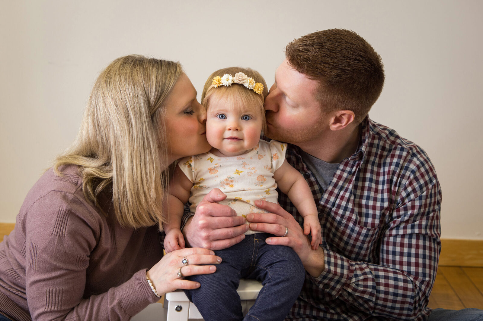 A couple holds their baby on a chair between them, each kissing one of her cheeks while she smiles at the camera.