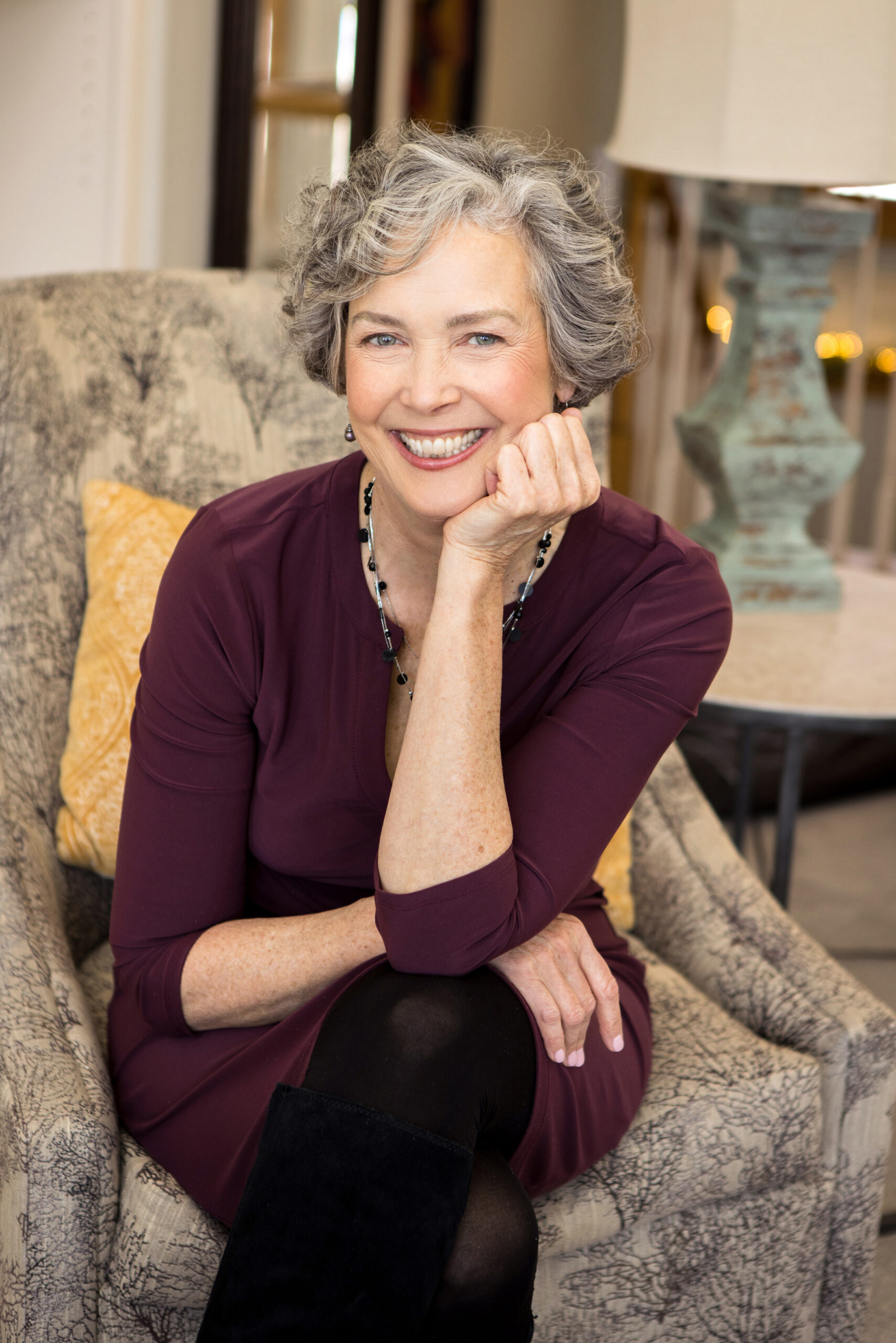 A professional heashot of a woman in a purple dress, posing on a chair. She is smiling brightly at the camera.