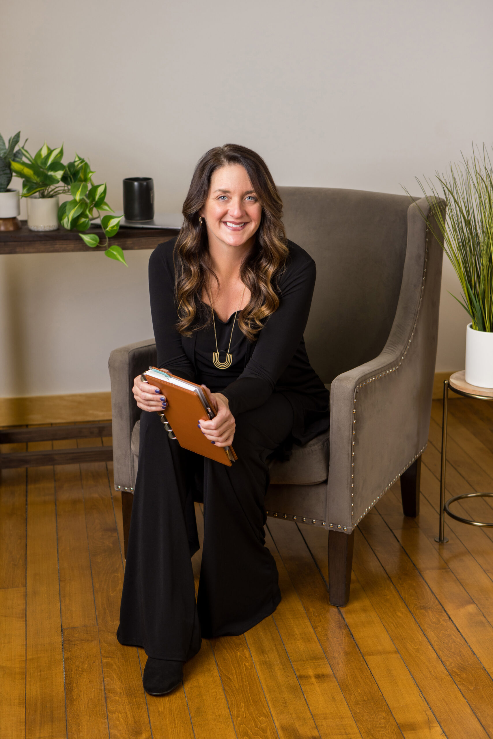 A professional photography session of a nicely dressed woman seated on a grey chair. She holds a brown journal or agenda style book.