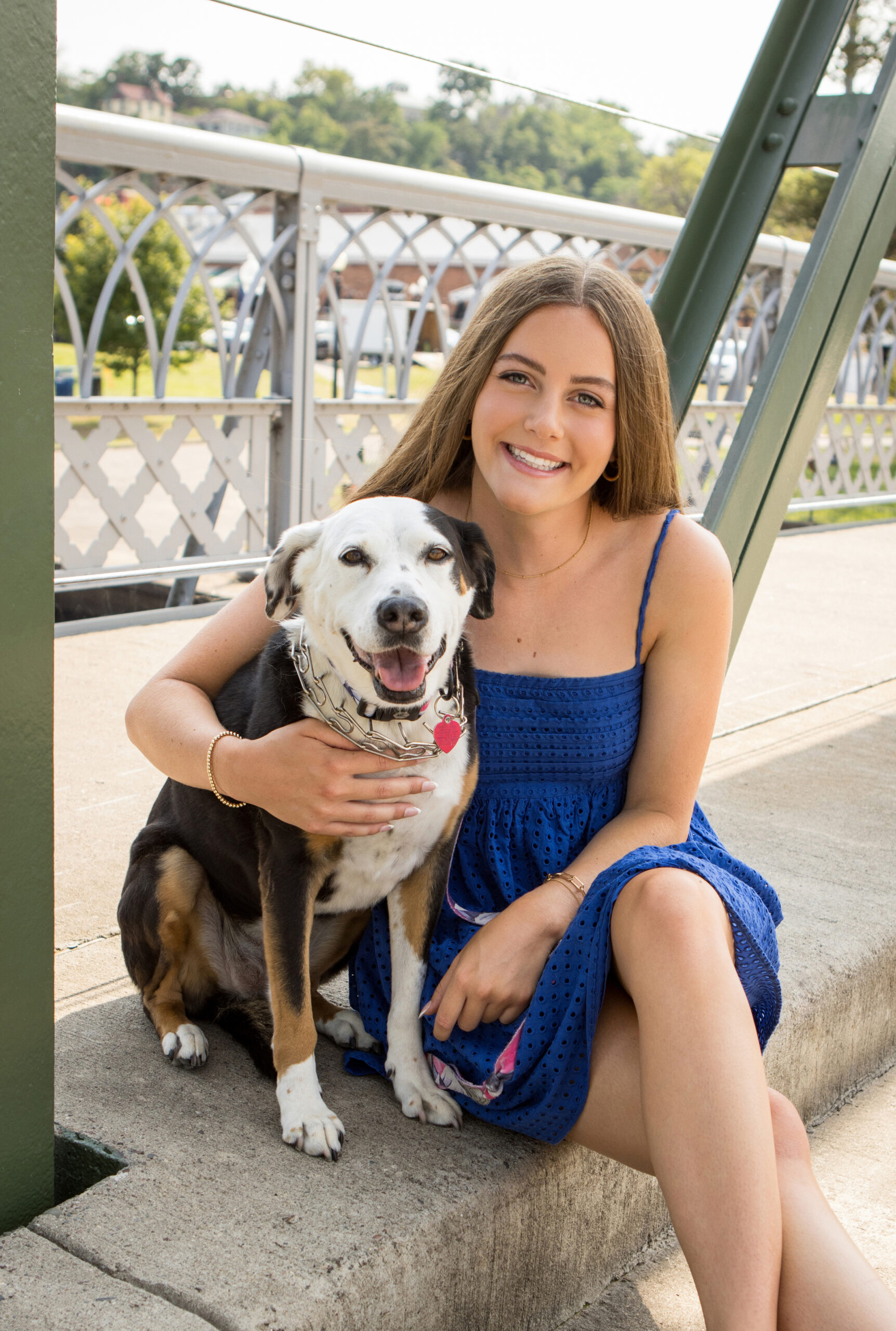 A professional photograph taken outside on a bridge of a High School Senior girl and her dog. She is wearing a blue dress and has one arm around her dog who is tri-colored with a short coat.