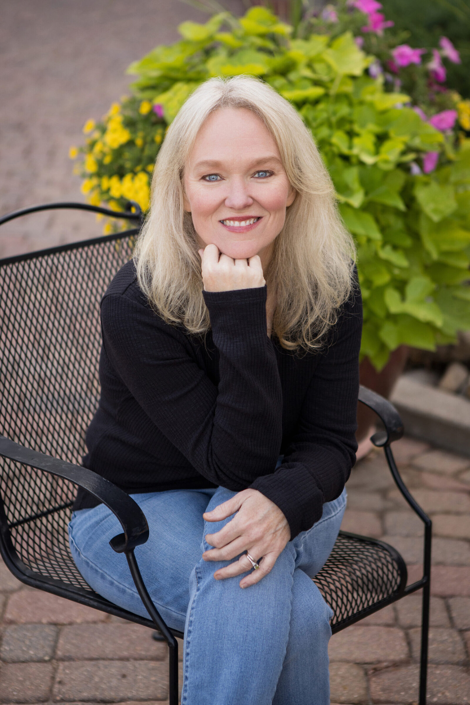 A professional headshot of a casually dressed woman sitting outside on a black bistro style chair.