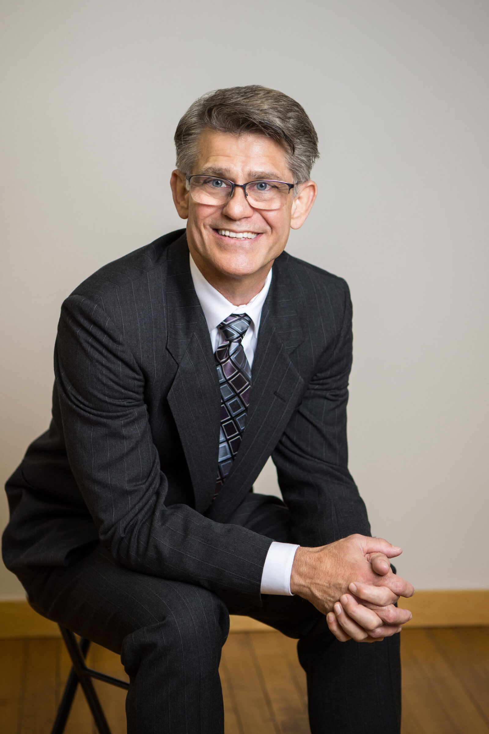 A man dressed in a nice business suit sits on a chair smiling at the camera for his in studio professional headshot session