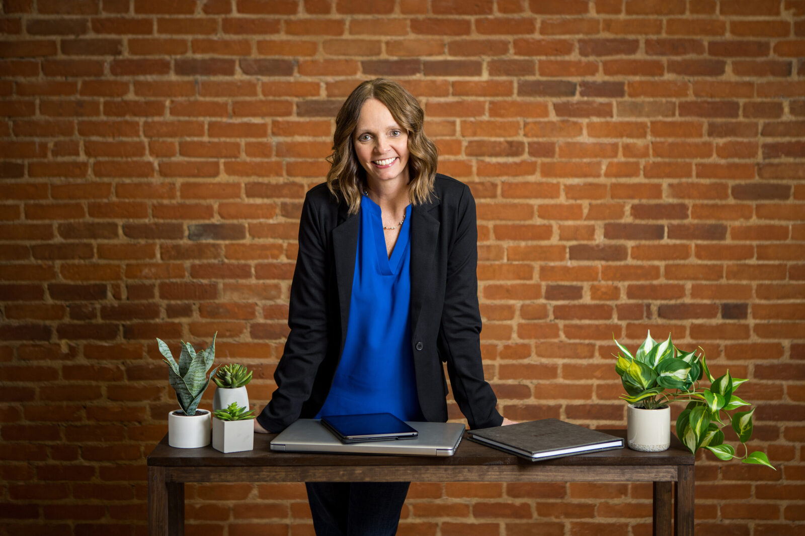 A business woman leans on her desk in front of a brick wall smiling at the camera for a professional business photo.