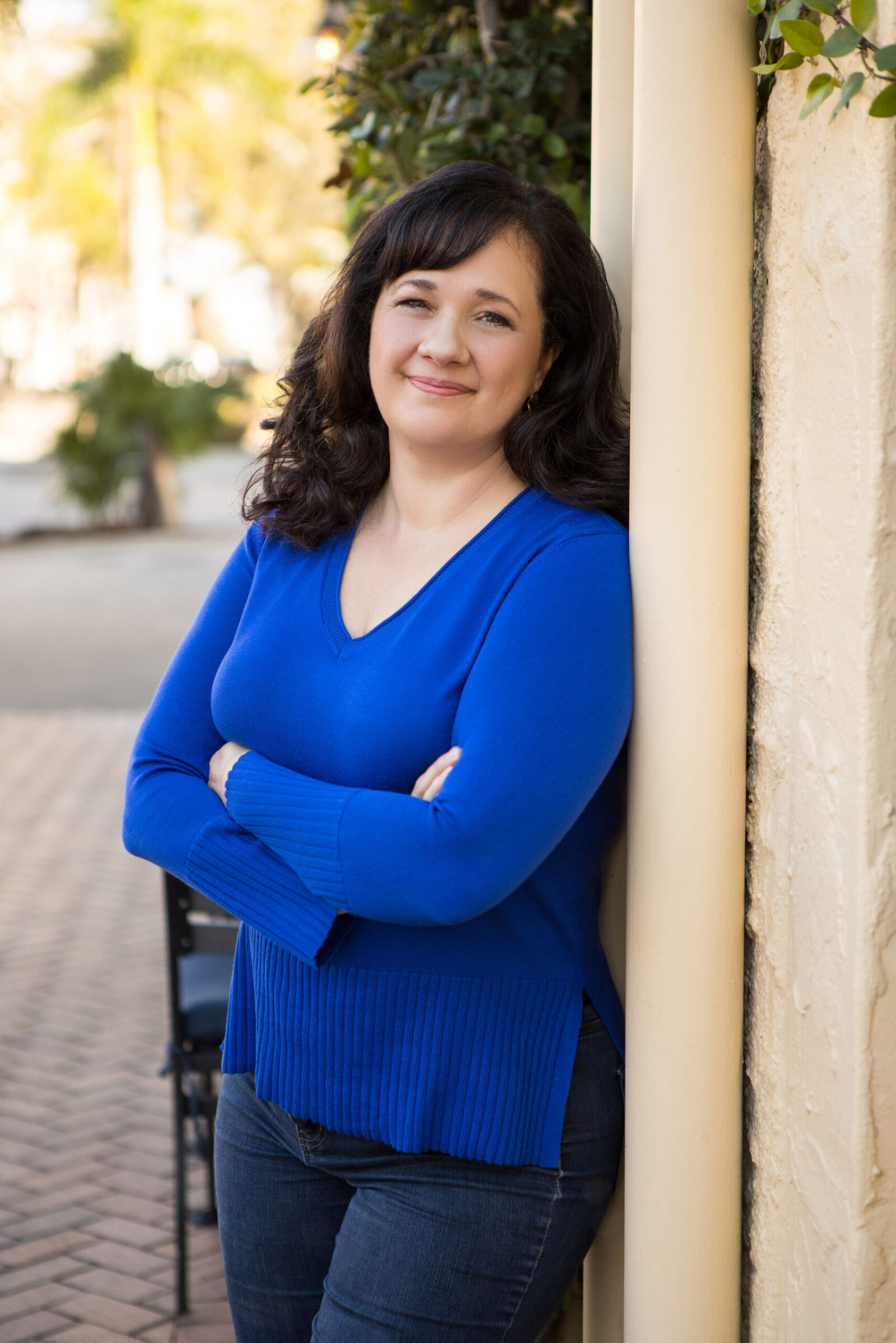 An outdoor professional headshot session for a business woman dressed casually in blue jeans and a blue top. She leans against a building with her arms crossed, smiling at the camera.