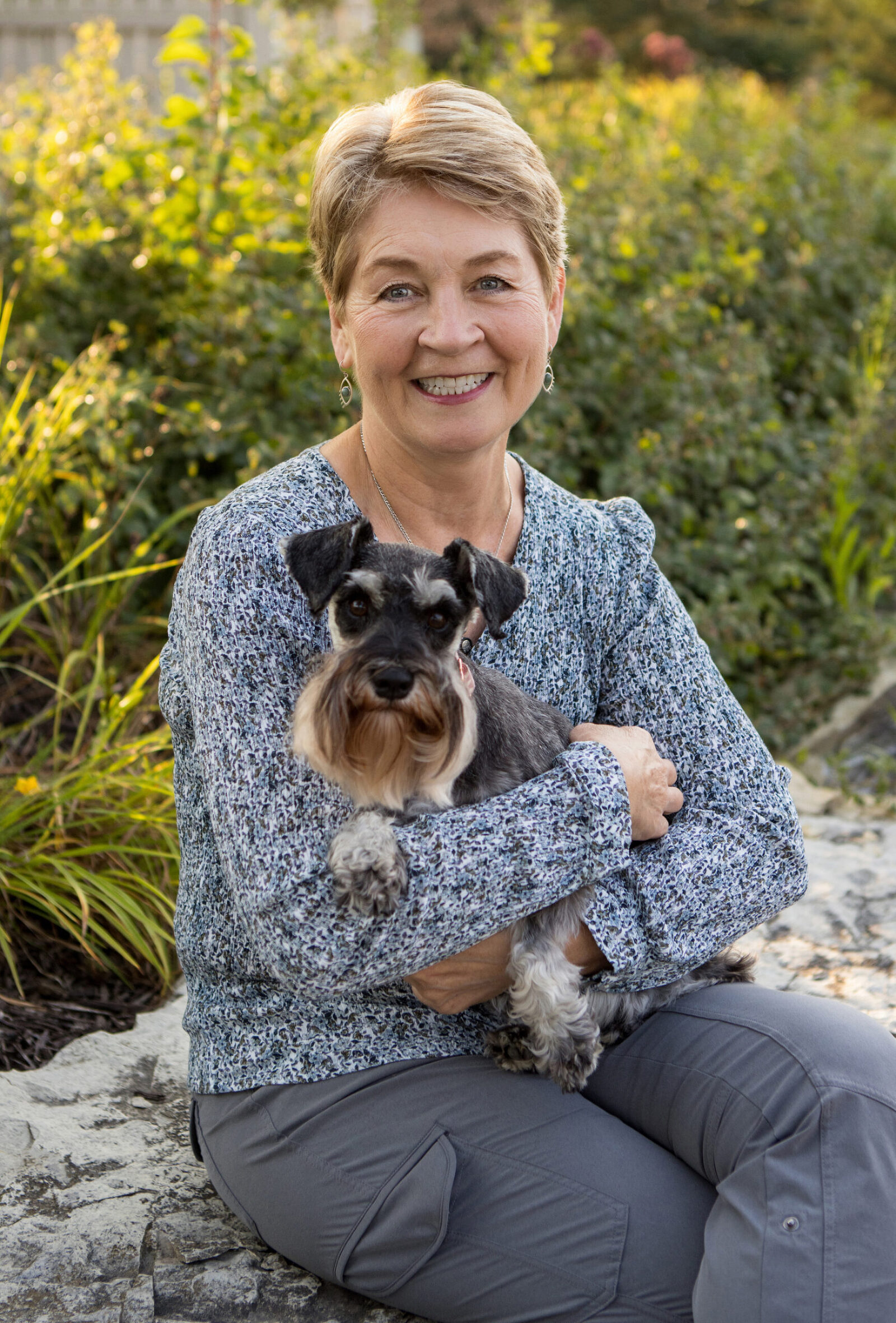 A nicely dressed woman sits on a boulder in a park with a big smile on her face. She is holding a small multicolored dog in her arms.