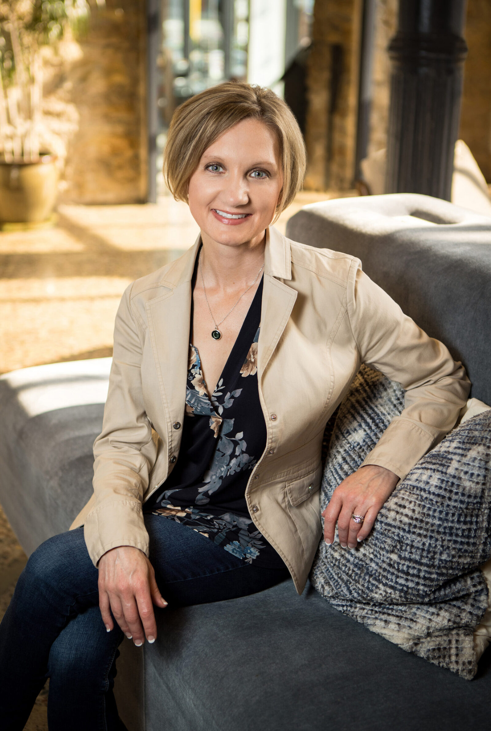 A professional headshot photo of a woman dressed in business casual attire sitting on a couch facing the camera to her left.
