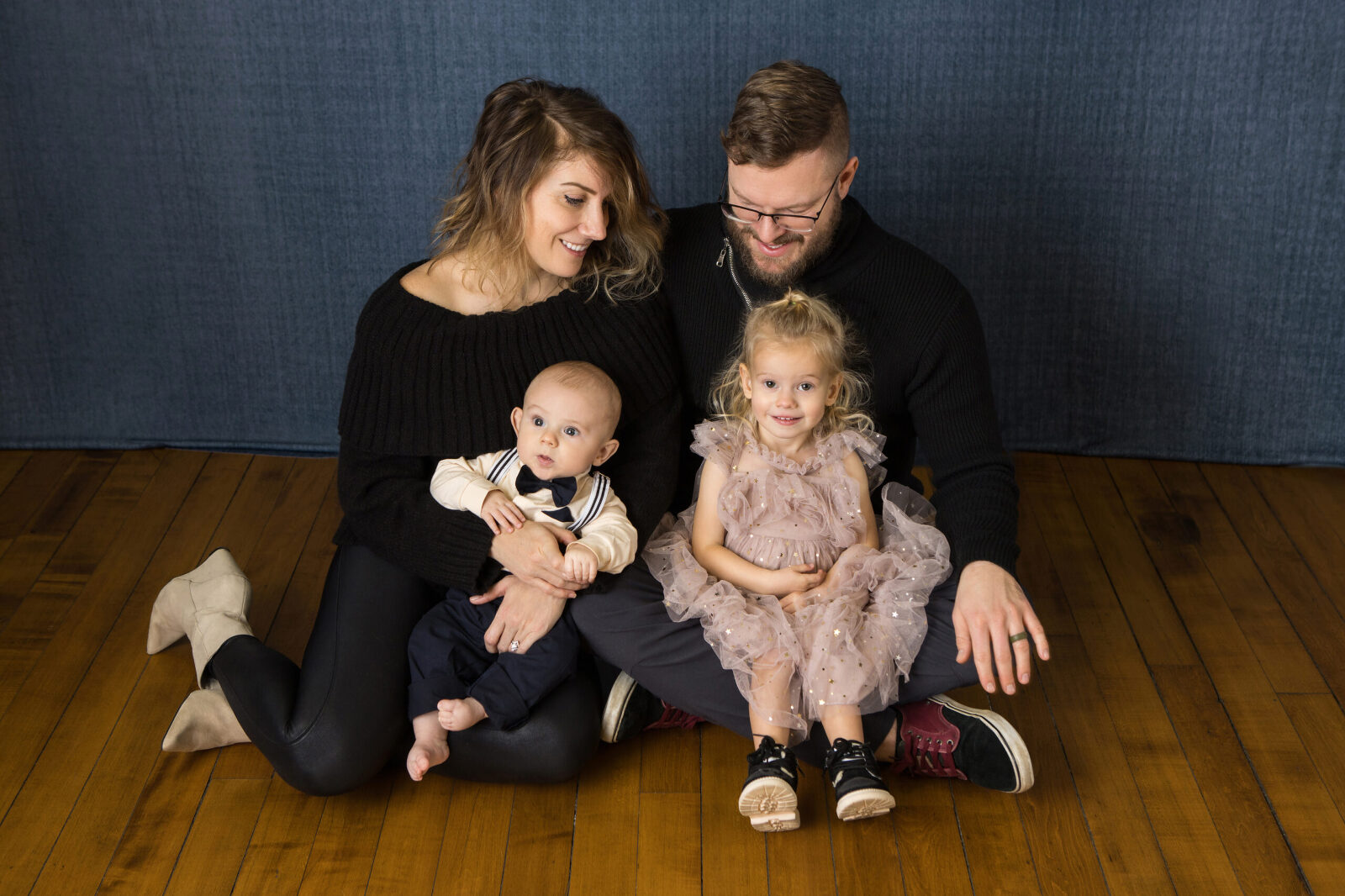a studio portrait of a family of four sitting casually on the floor. The mom and dad each have a child snuggled on their laps. The older child is a young girl in a pink dress and the baby is a boy wearing a bow tie and suspenders.