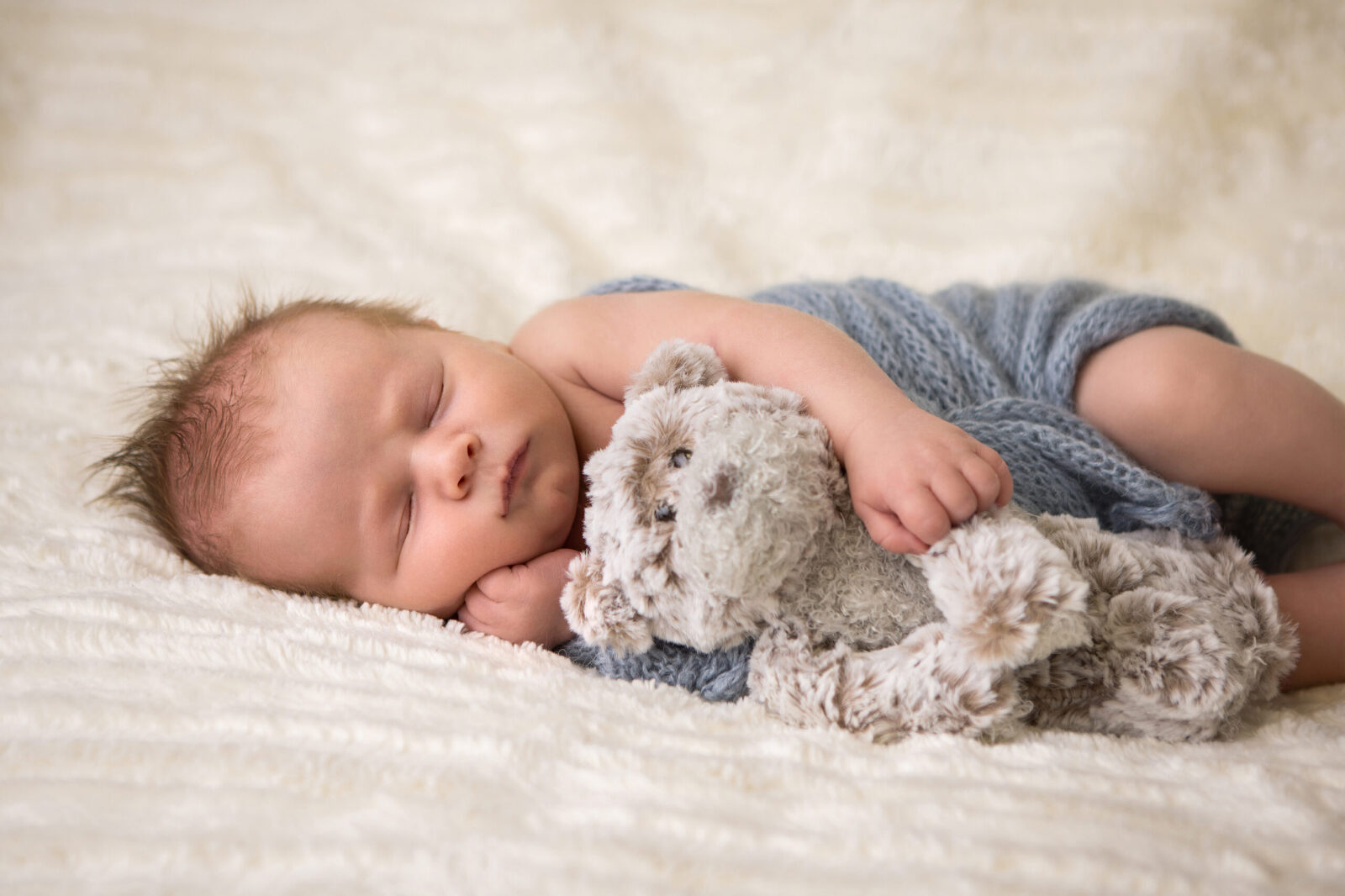 An infant sleeps on their side with one arm under his cheek and the other snuggling a teddy bear. He is swaddled in a blue knit blanket.