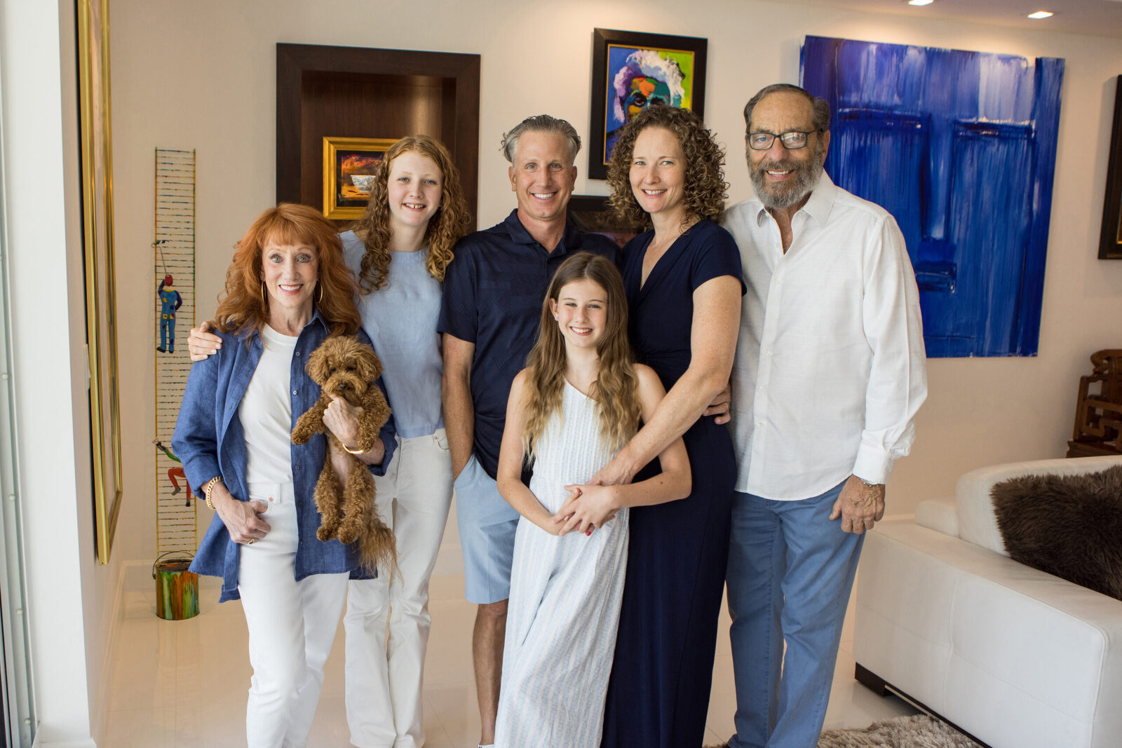 A family of 6, plus their little dog, stand together in a brightly lit room smiling for a family portrait. They are all coordinated in blue's and whites which matches the aesthetic of the room they are in
