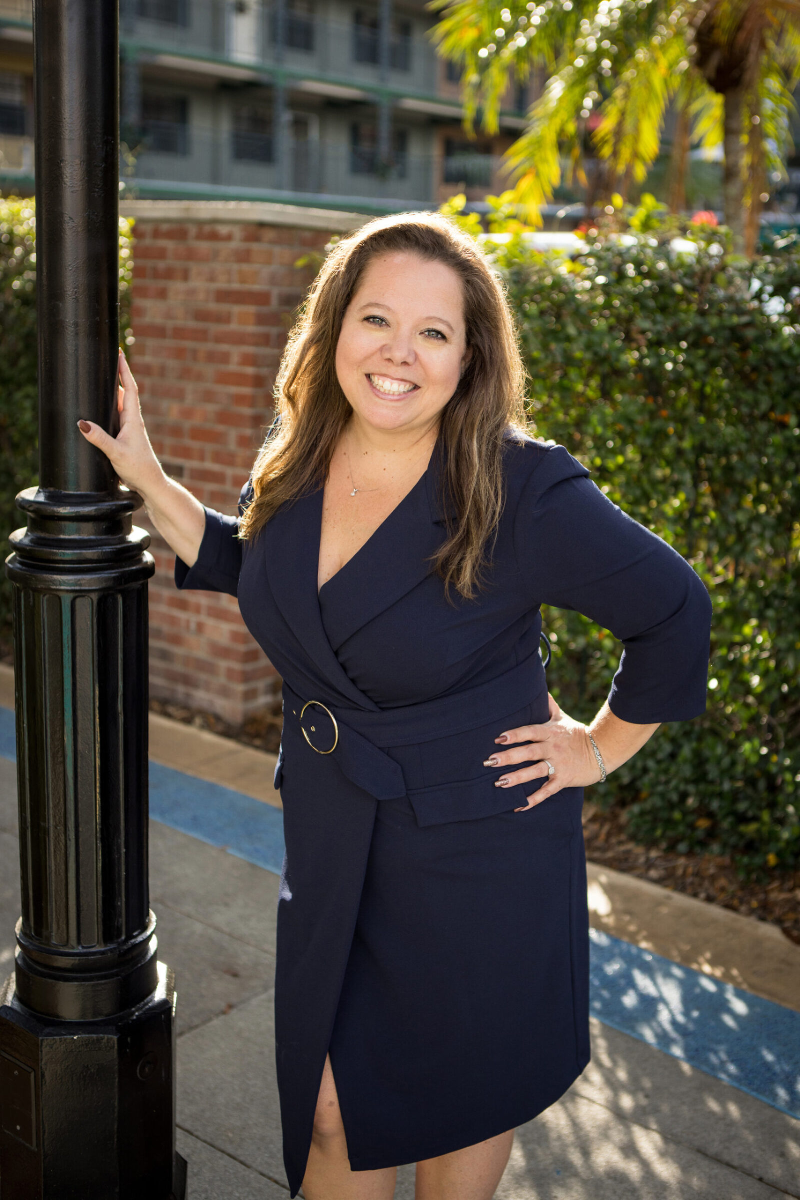 A woman poses outside near a lightpole in front of a motel. She smiles brightly at the camera, wearing a dark business dress.