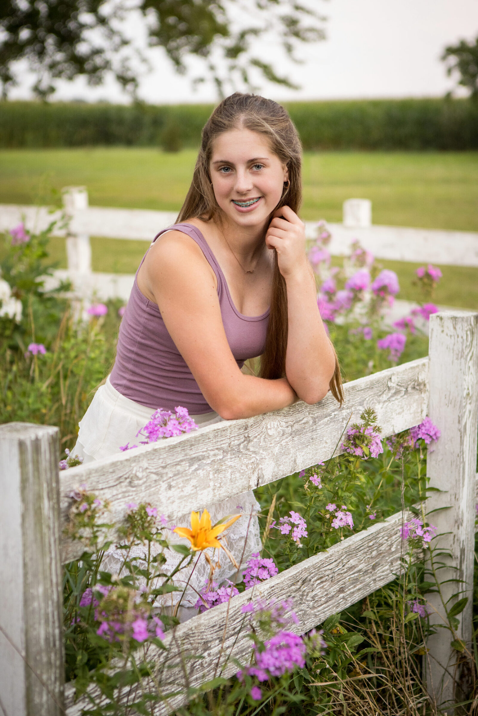 A young girl is smiling at the camera as she leans againt a white fence in a pasture of flowers. She is wearing a white flowy skirt and a purple tanktop. Her pose is styled but still natural.