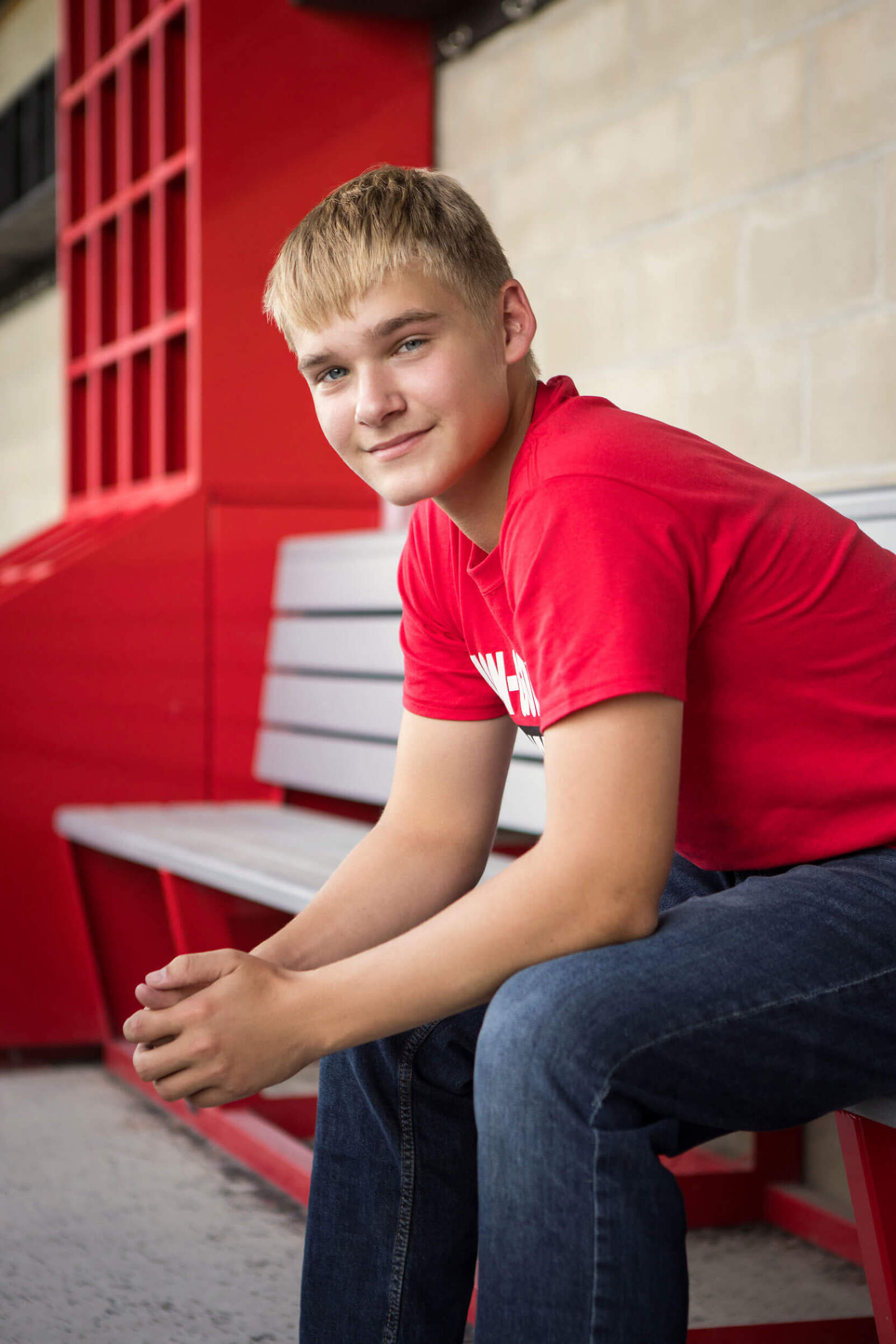 A high school senior boy sits in a baseball dugout smiling at the camera. He is wearing jeans and red school mascot t shirt which matches the accent color in the dugout.