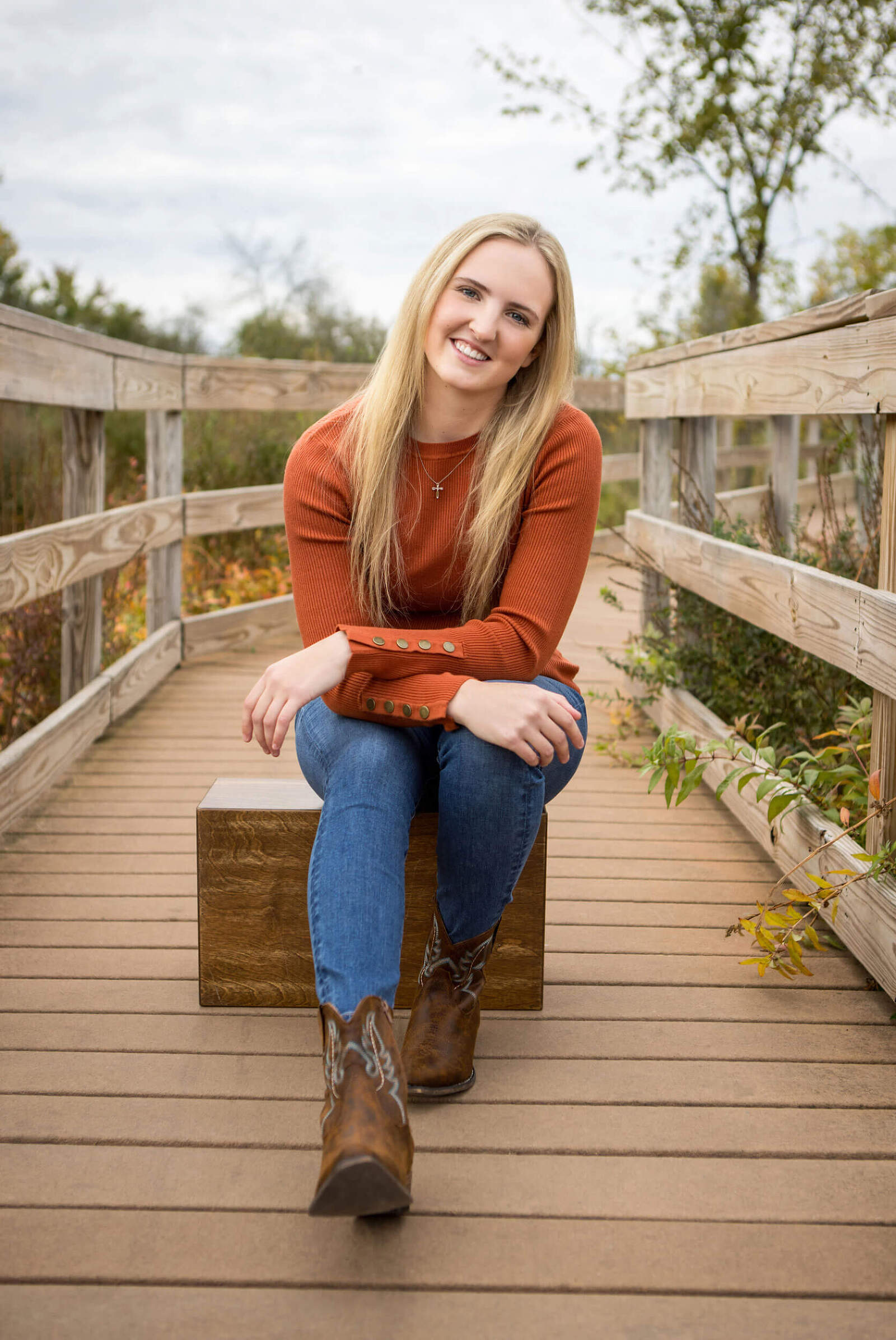A senior picture of a young woman sitting in the middle of a small foot bridge. She is wearing jeans and cowboy boots with a burnt orange top. She is in a relaxed, seated pose and smiling at the camera.