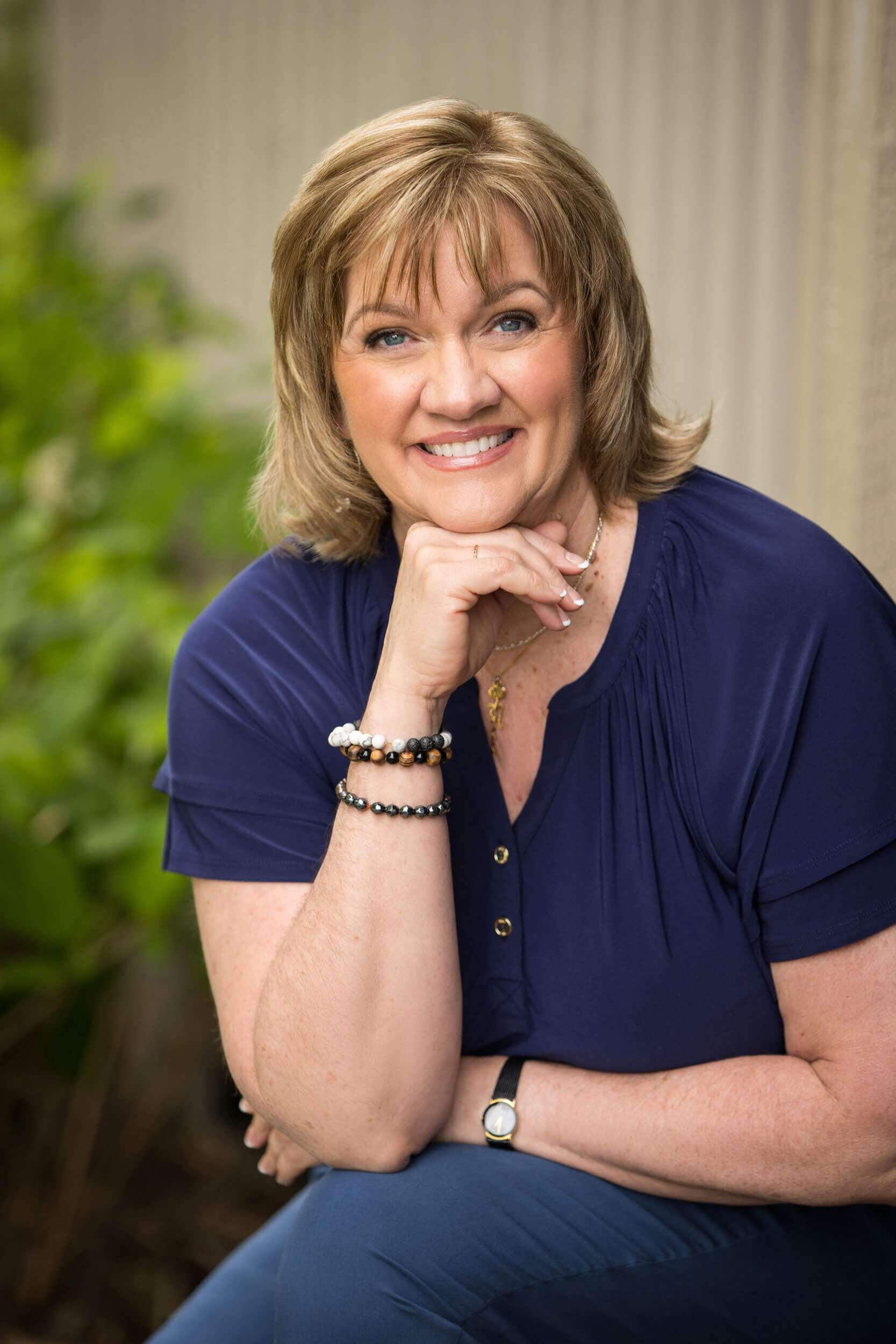 A professional headshot of a woman dressed in a navy top and pants. She is seated cross-legged, resting her chin on her hand. She is smiling face on to the camera.
