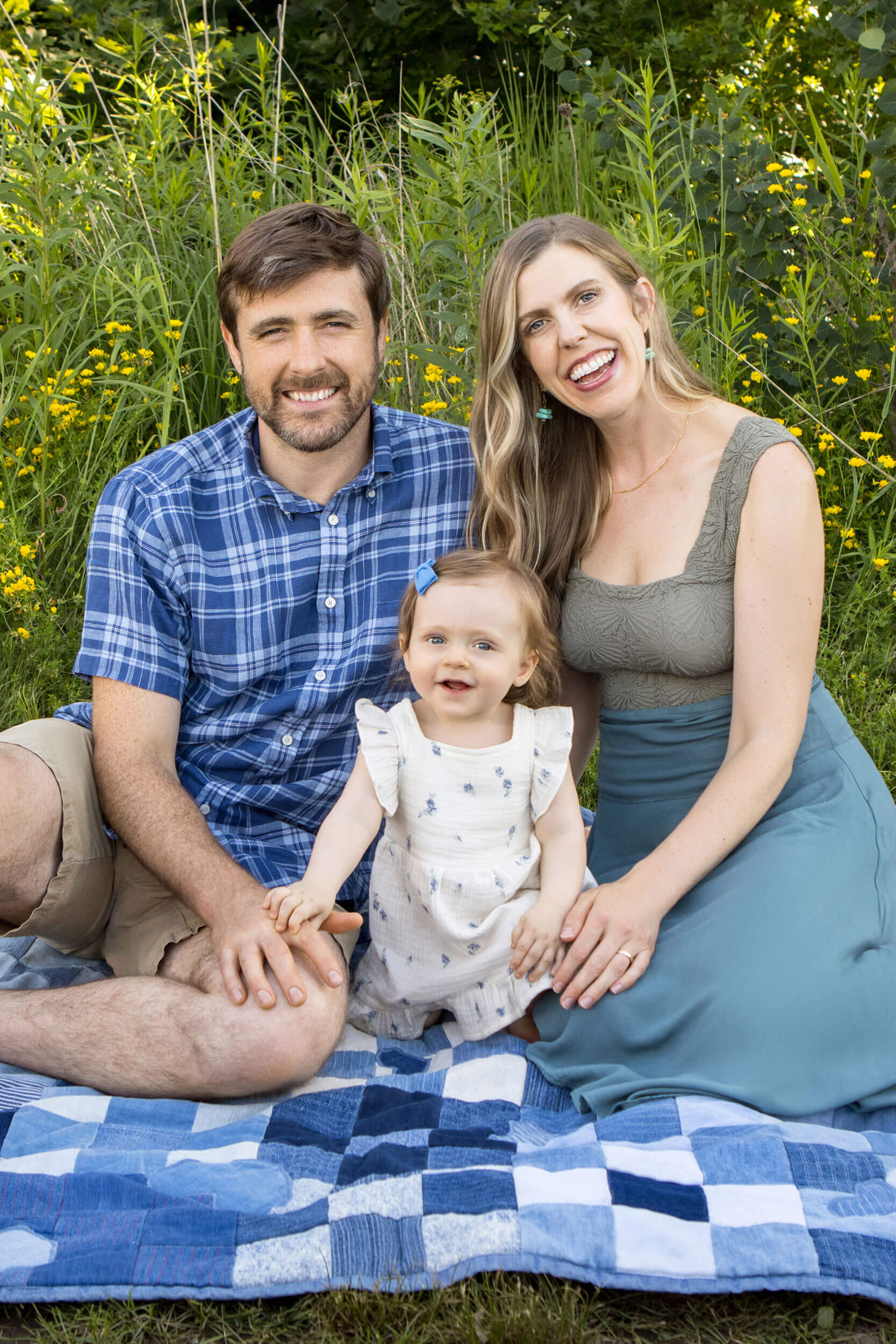 A young couple and their toddler pose on a quilt in a park for their family photography session with Kristina
